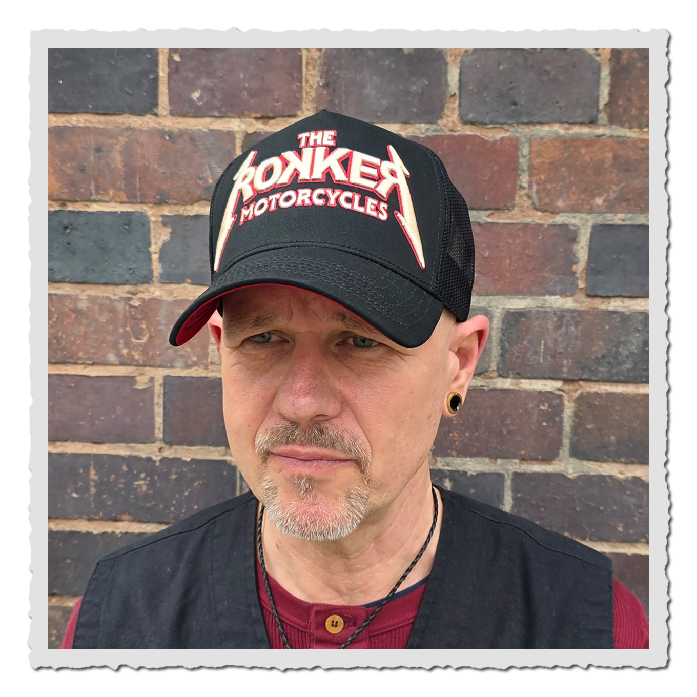 Mann vor Ziegelmauer trägt schwarz-rote Trucker-Cap mit gesticktem Schriftzug, schräg von vorn fotografiert.